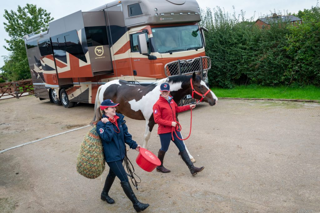 Riders leading horse with haynet and feed bucket, with horsebox in the background